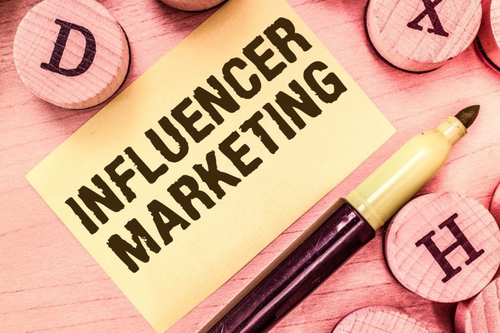 A close-up shot of a sticky note with the bold text "INFLUENCER MARKETING" written on it, placed on a wooden surface next to a highlighter pen and wooden circular tiles.