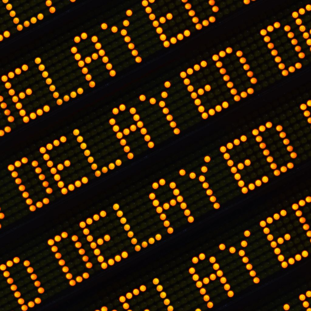 A close-up, diagonal view of a digital display board showing the word "DELAYED" repeated in glowing orange dot-matrix text.