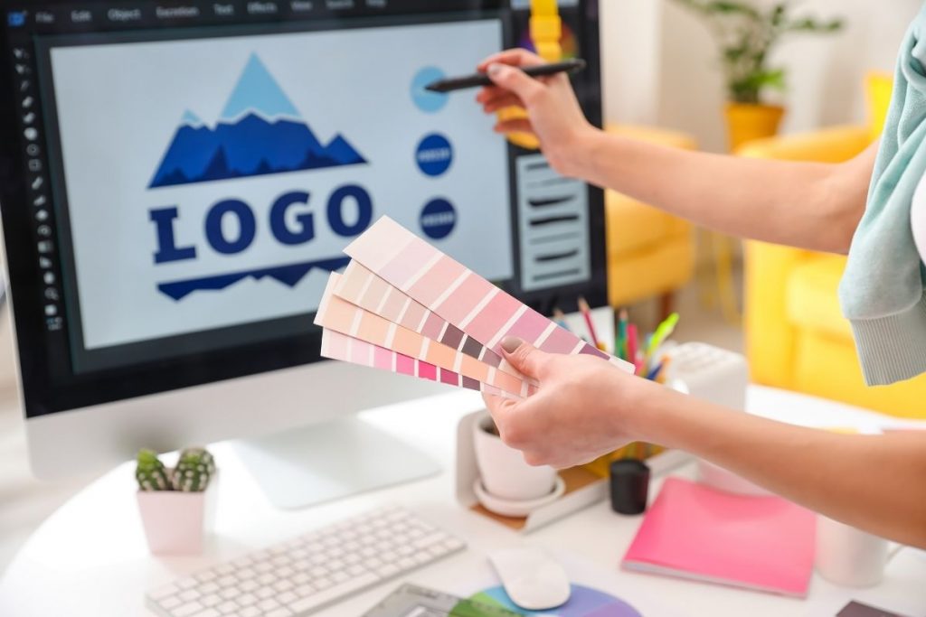 A designer working on a computer screen showing a blue mountain logo design, while holding up a fan of colour palette swatches for selection.