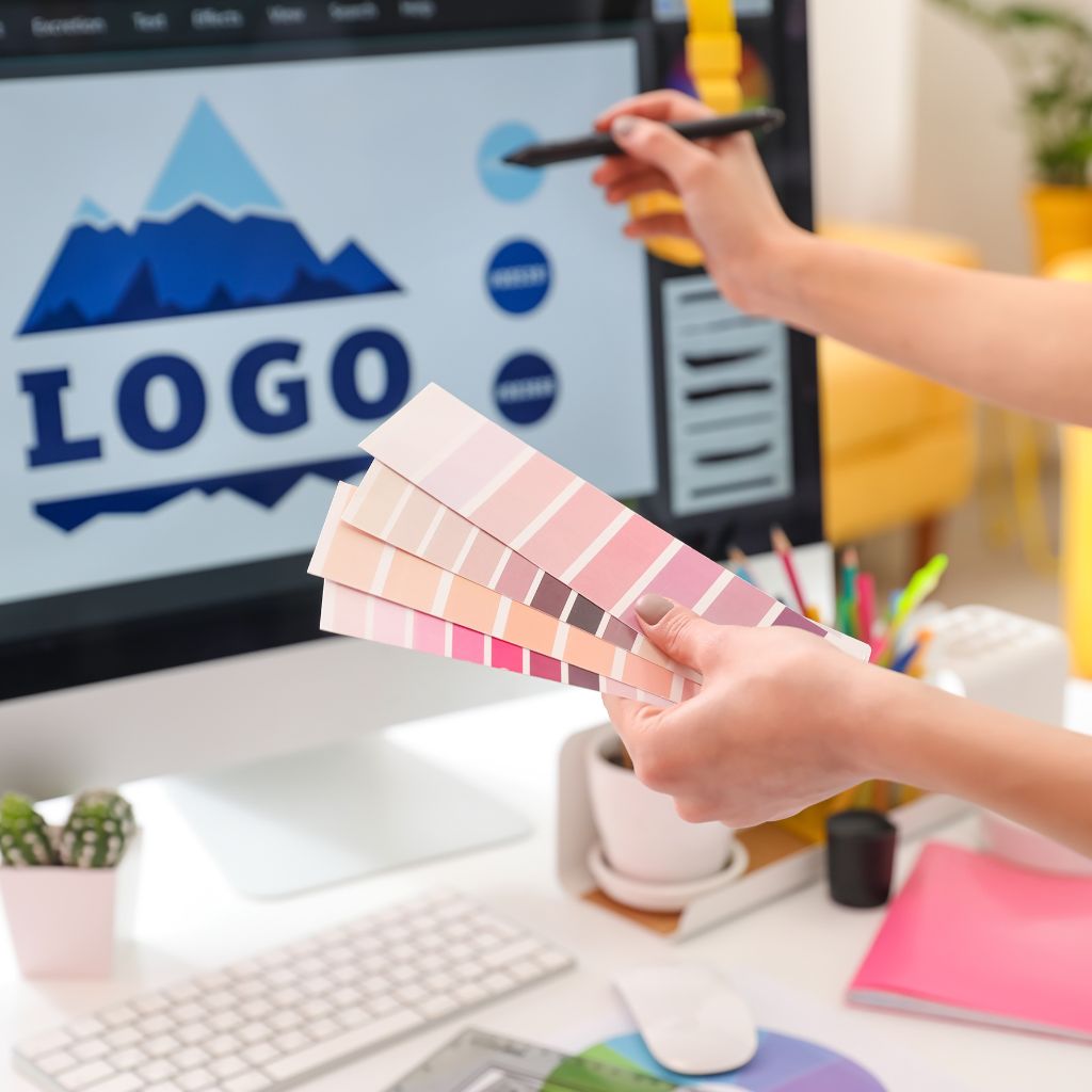 A designer working on a computer screen showing a blue mountain logo design, while holding up a fan of colour palette swatches for selection.