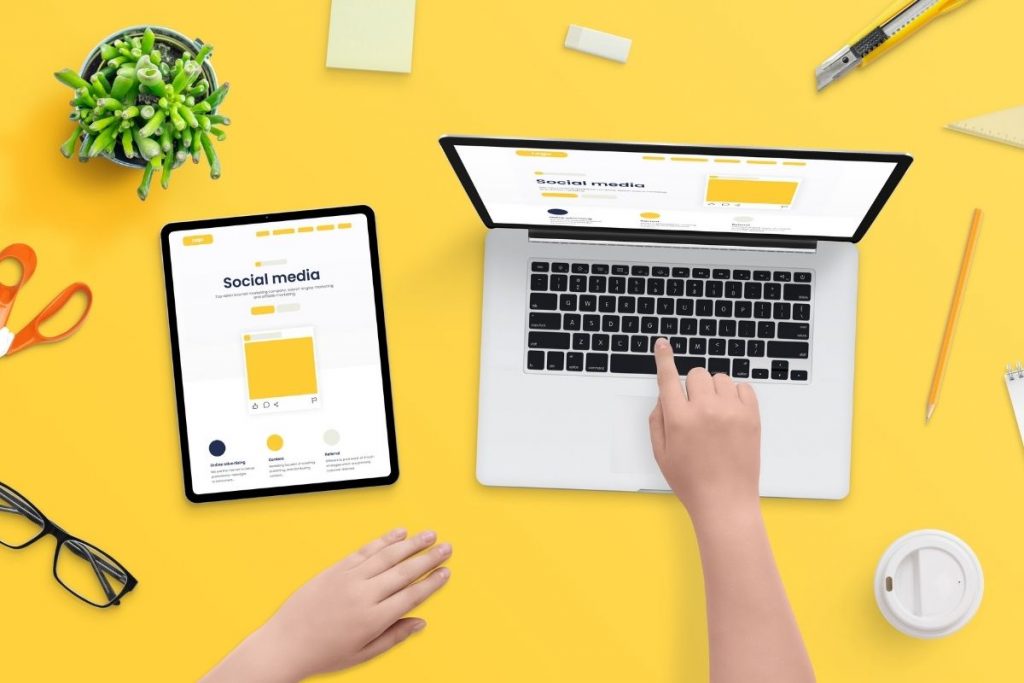 An overhead desk view showing a hand touching a laptop keyboard, next to a tablet displaying a "Social media" graphic, all on a bright yellow background.