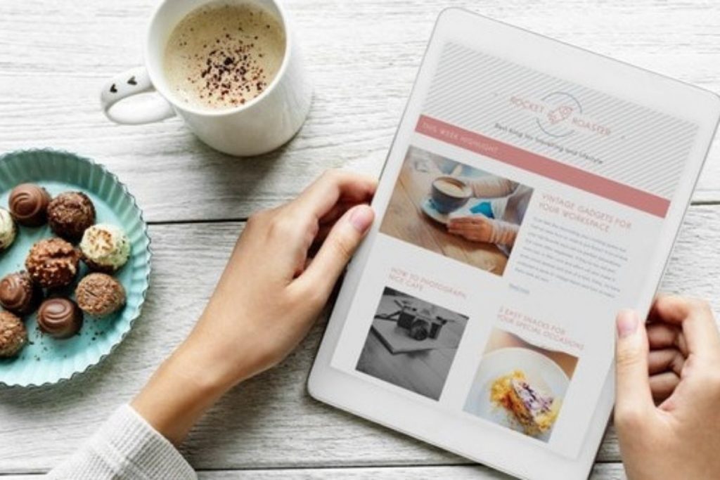An overhead shot of hands holding a tablet displaying a blog post on a white wooden table, next to a mug of coffee and a plate of chocolates.