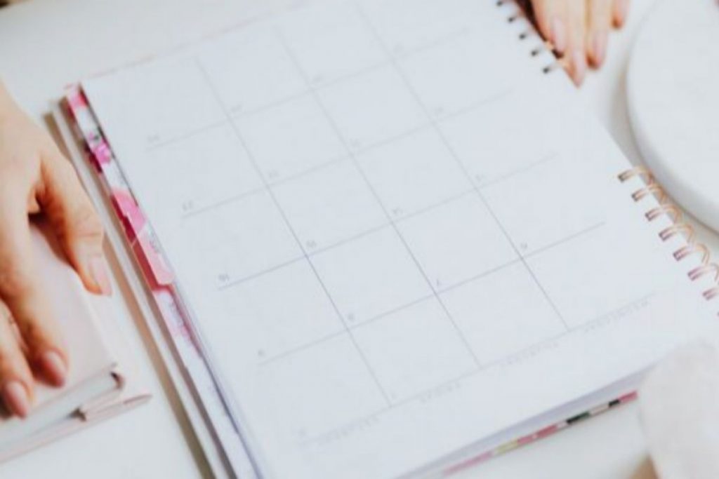 A pair of hands resting on an open weekly calendar or planner, with a pink pen, spool of yarn, and jewellery accessories scattered nearby on a white desk.