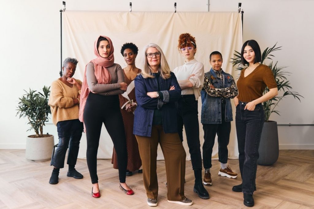 A diverse group of six women of different ages and backgrounds standing together in a studio setting in front of a neutral backdrop, looking confidently at the camera. They are posed with their arms crossed or hands on their hips, representing International Women's Day.