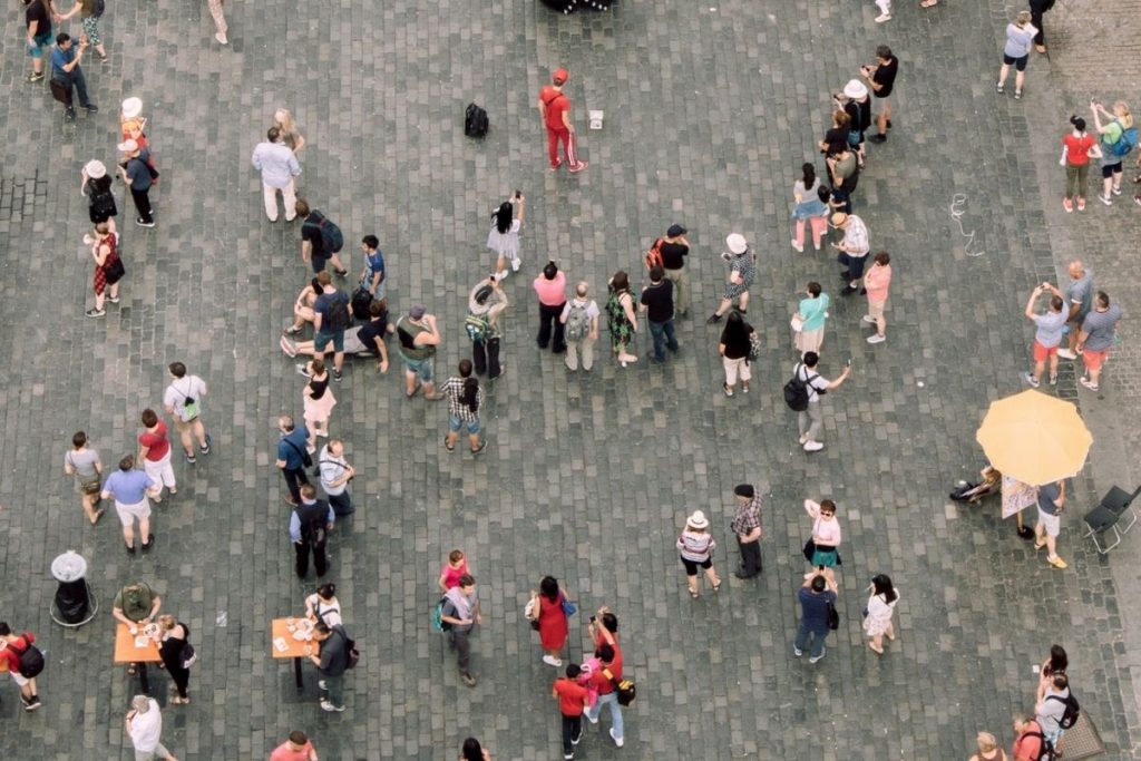 High-angle aerial shot of a large group of people scattered across a dark gray cobblestone square, with one person in a distinctive bright red outfit standing out near the centre, symbolising standing out in a crowded market.