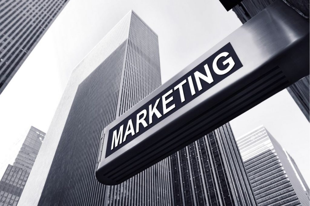 A black and white, low-angle photograph of tall city skyscrapers, with a large, angled metallic sign bearing the capitalised white word "MARKETING" silhouetted against the bright sky.