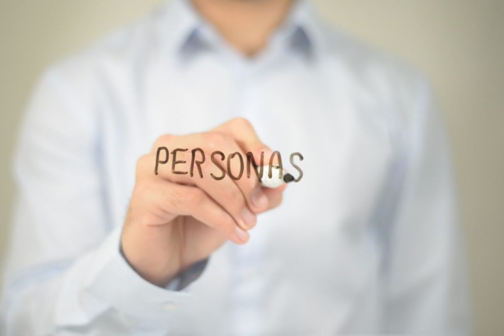 A blurred person in a light shirt writes the word "PERSONAS" in black marker directly onto a transparent glass or screen.