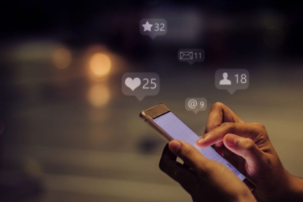 Hands holding a smartphone in a dark setting, with fingers interacting with the screen, surrounded by floating social media notification icons for likes, followers, messages, and mentions.