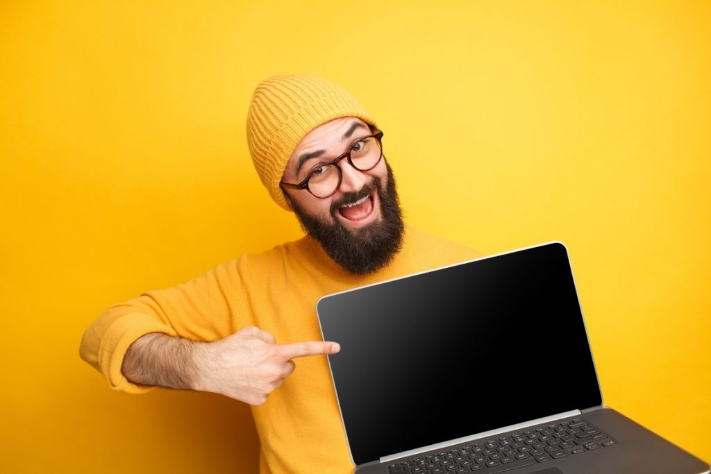 A cheerful, bearded man in a yellow knit hat and yellow sweater smiles broadly, pointing to the blank black screen of an open laptop, set against a solid yellow background.