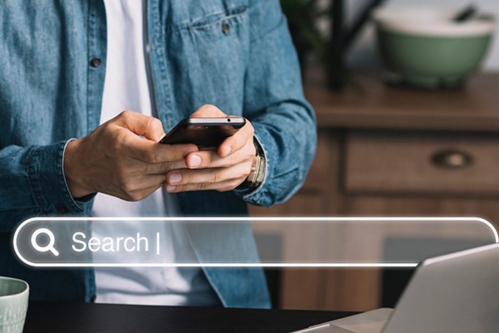 A person in a denim shirt holds a smartphone over a desk with a laptop, with a large, glowing "Search" bar overlayed across the foreground.