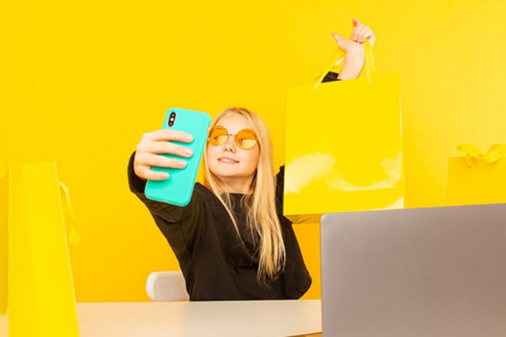 Young woman with sunglasses taking a selfie with a light blue smartphone while holding up a bright yellow shopping bag, against a yellow background.