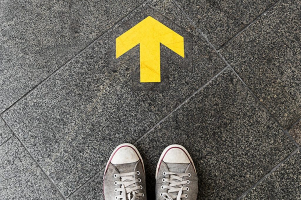Overhead view of a person's feet in gray sneakers standing on dark gray tiled flooring, with a bright yellow arrow painted on the floor pointing forward.