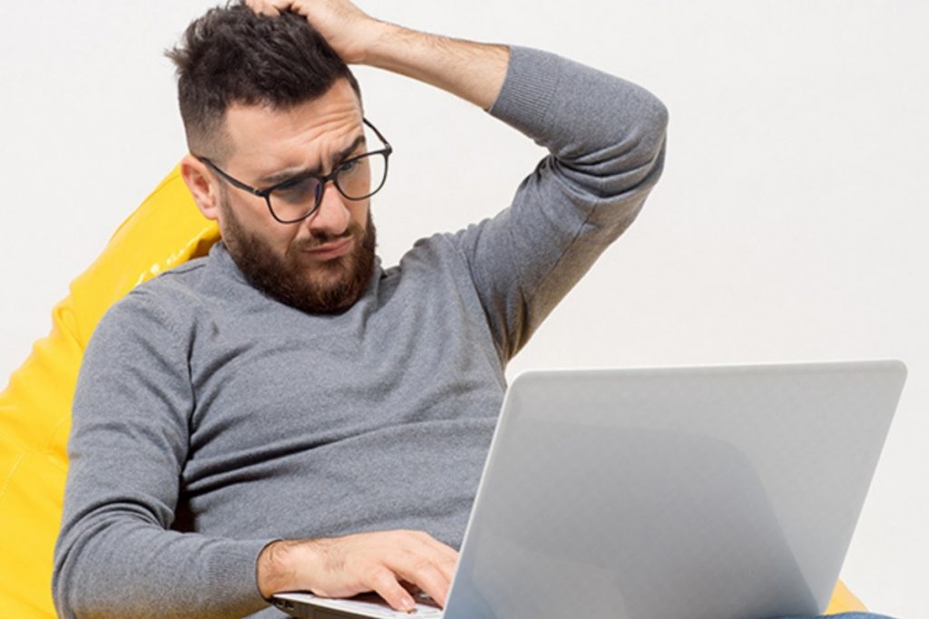 A bearded man wearing glasses and a gray sweater sits in a yellow beanbag chair, looking confused while working on a silver laptop.