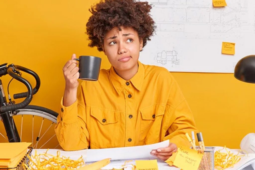 A woman with curly hair sitting at a desk with a yellow background, holding a coffee mug and looking thoughtfully at a document.