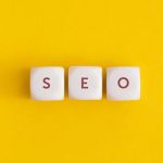 Three white cubes placed in a row against a solid yellow background, with the letters "S," "E," and "O" printed on them in red.