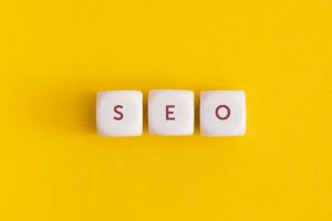 Three white cubes placed in a row against a solid yellow background, with the letters "S," "E," and "O" printed on them in red.