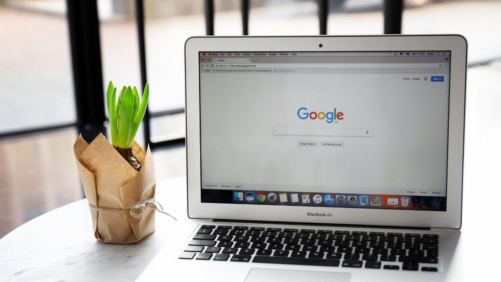 A MacBook Air laptop open to the Google search page, with a potted green plant wrapped in brown paper sitting next to it on a white table.