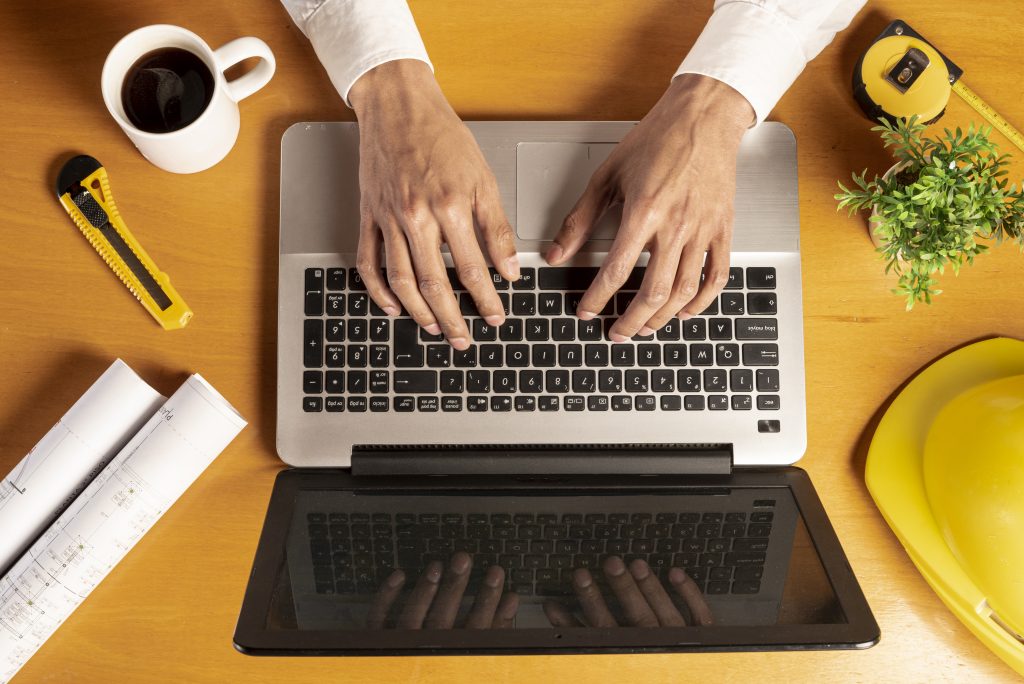 A top-down view of hands typing on a laptop keyboard. Surrounding the laptop are architectural blueprints, a yellow utility knife, a white mug of black coffee, a measuring tape, a small green plant, and a yellow construction hard hat, all resting on a wooden desk.