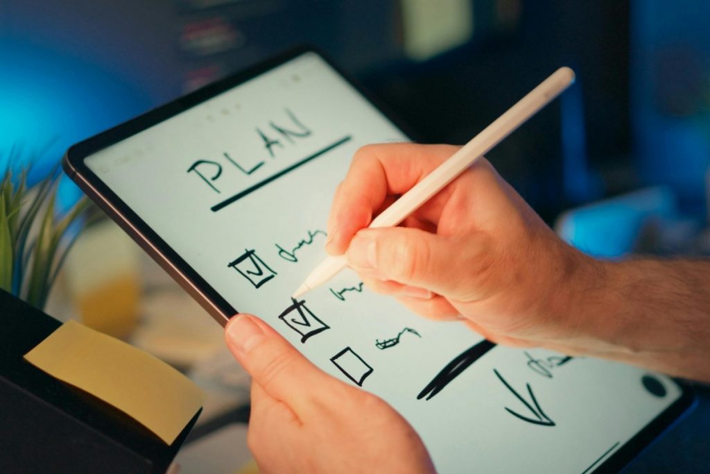 A person writing the word 'PLAN' and checking boxes on a digital tablet with a stylus.