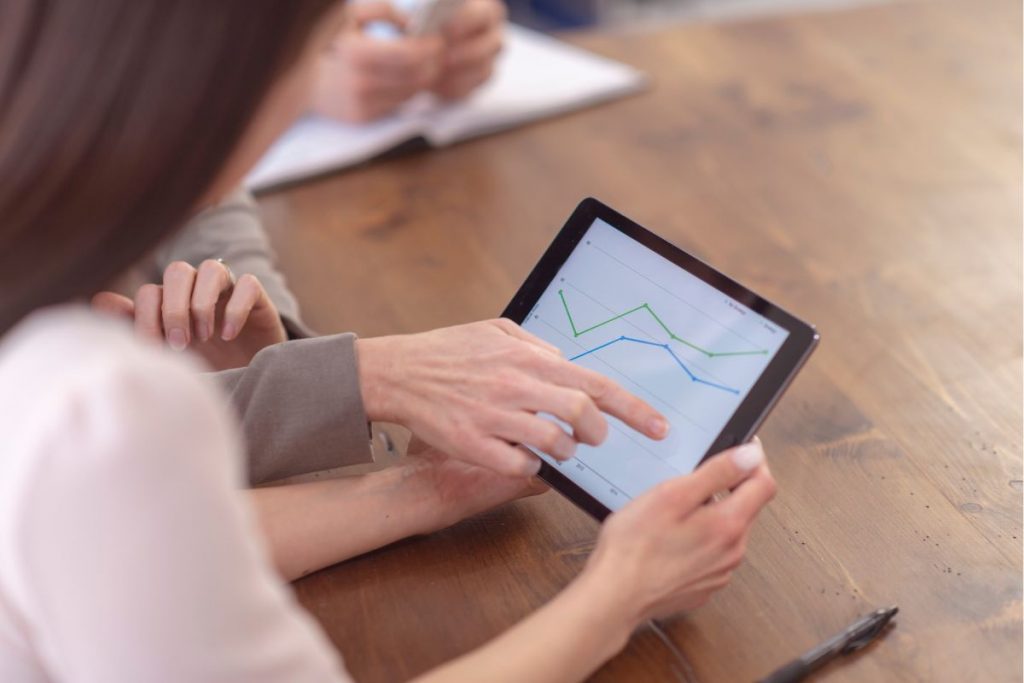 A close-up, over-the-shoulder shot of two people seated at a wooden table, one pointing to a digital line graph on a tablet screen that shows increasing and decreasing performance metrics.