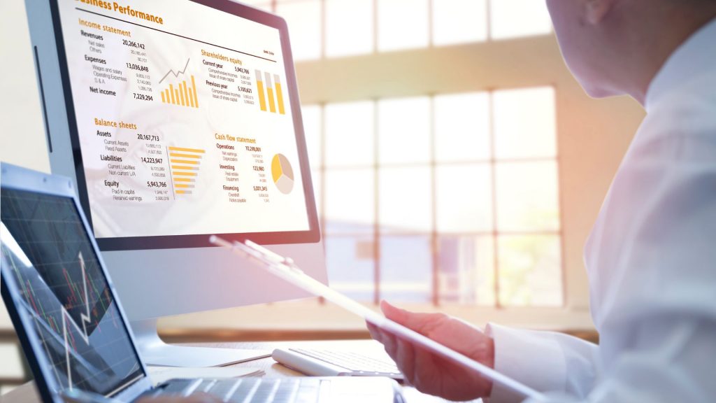 Computer monitor displaying a business performance dashboard with financial statements (Income Statement, Balance Sheets, Cash Flow Statement). A person is looking at a paper report.