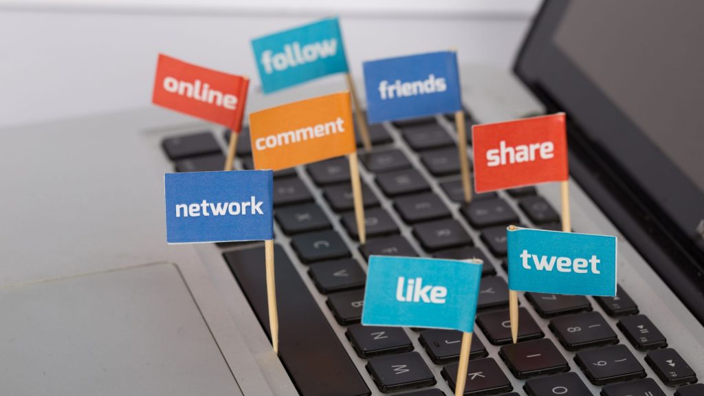Small flag signs with social media engagement words (like, share, follow, comment, tweet, network, friends, online) stuck in a laptop keyboard.