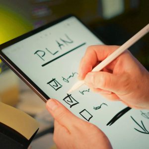 A person writing the word 'PLAN' and checking boxes on a digital tablet with a stylus.