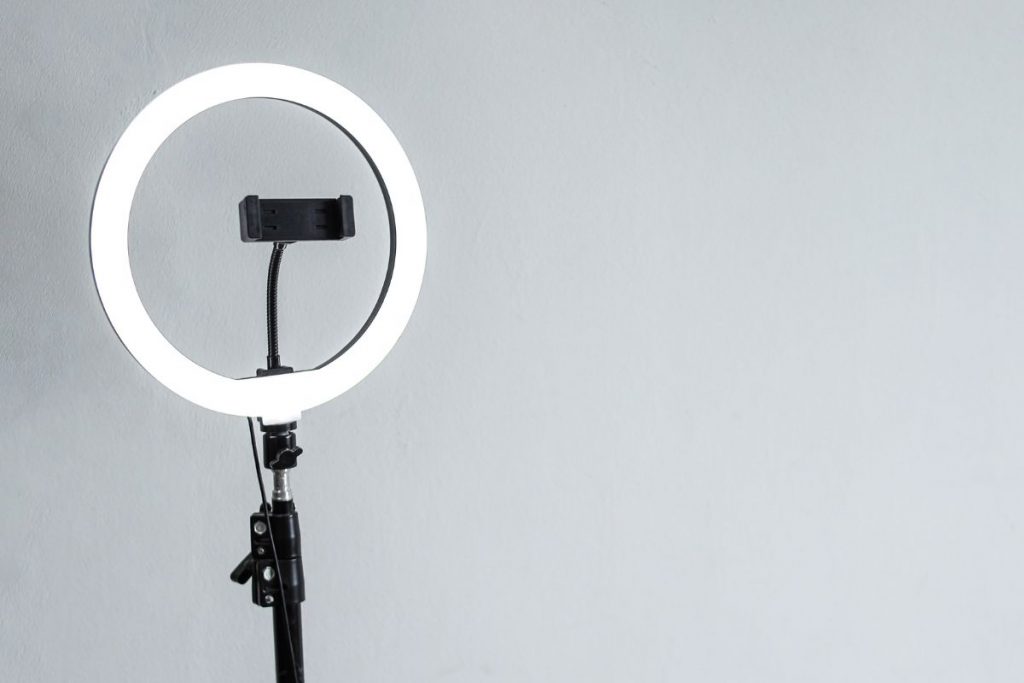 A professional ring light on a stand, with a smartphone holder mounted inside, set against a plain light gray wall, used for filming video content.