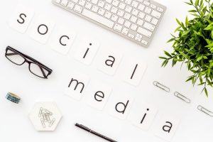 A flat lay of a desk with the words "SOCIAL MEDIA" spelled out using individual white cards with black letters. The arrangement includes a keyboard, eyeglasses, paperclips, a pen, and a small green plant on a white background, symbolising social media marketing.