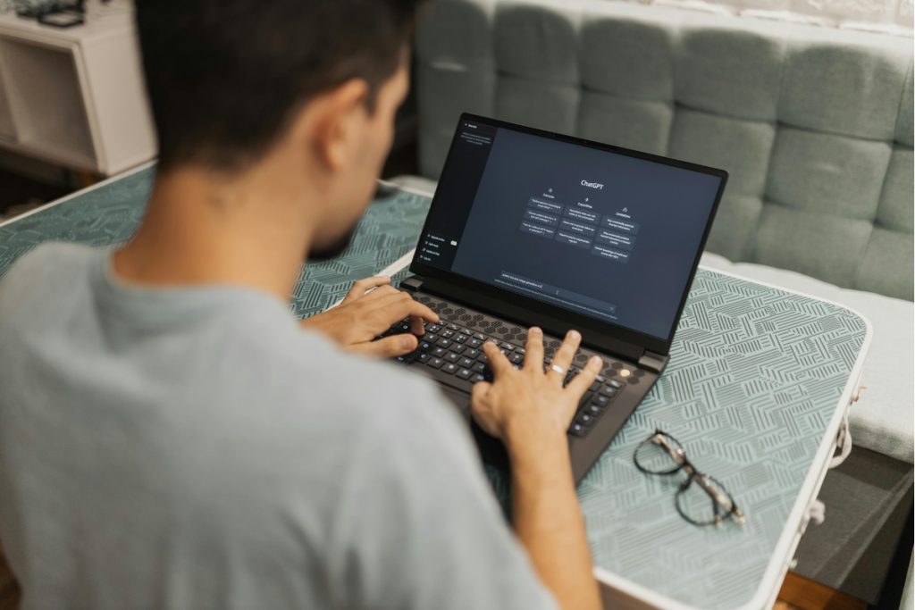 Over the shoulder shot of a man typing on a laptop, with the ChatGPT interface visible on the screen, sitting on a patterned surface.