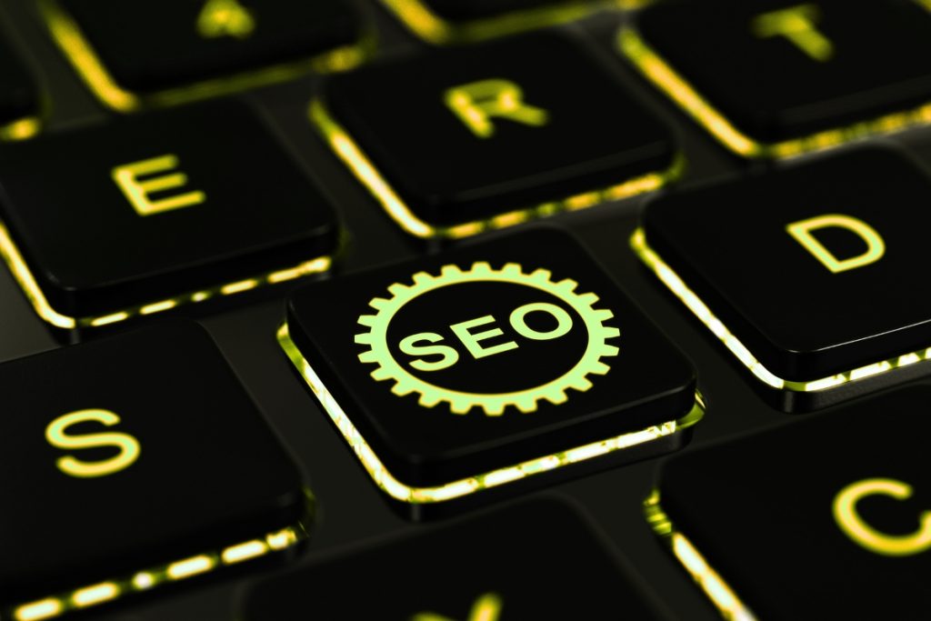 A close-up of a dark keyboard with back-lit keys, featuring one key with a glowing yellow-green gear icon and the letters "SEO" in the centre.
