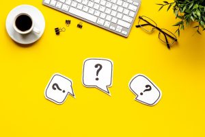 A top-down view of a yellow desk with a keyboard, coffee cup, glasses, and three paper speech bubbles containing question marks.