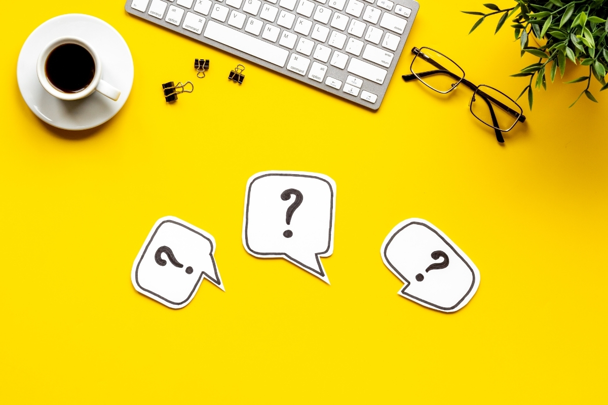 A top-down view of a yellow desk with a keyboard, coffee cup, glasses, and three paper speech bubbles containing question marks.
