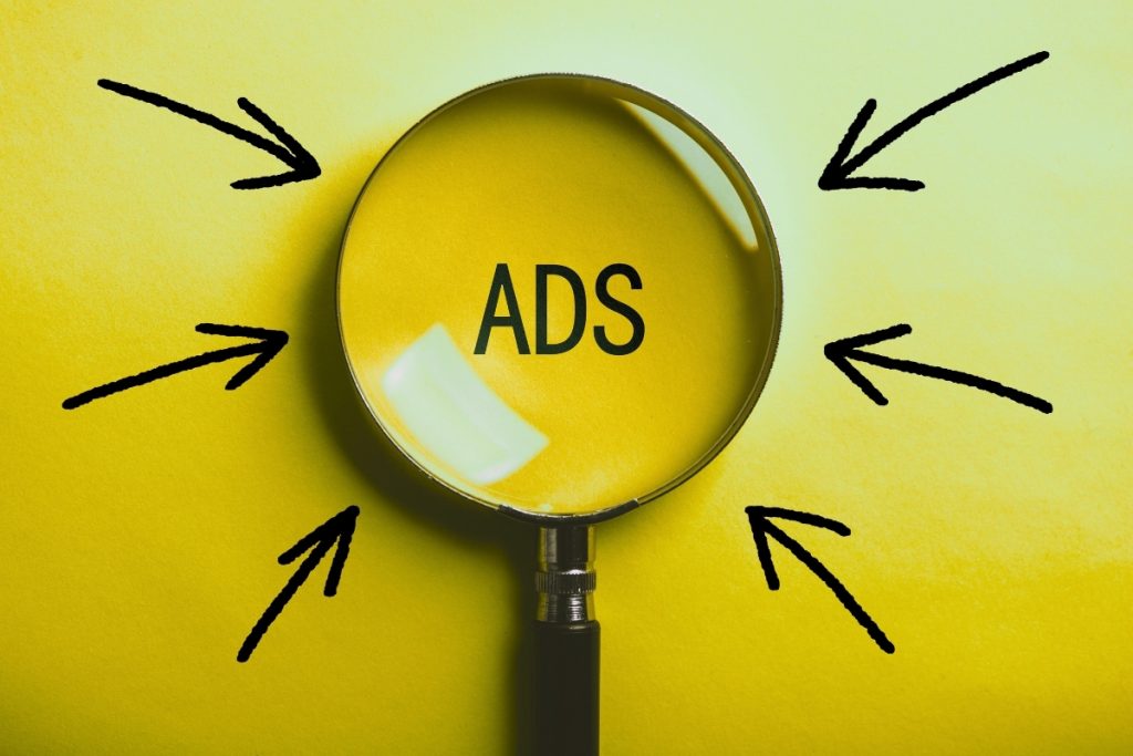 A magnifying glass centered over the word "ADS" on a yellow background, with black arrows pointing toward the lens.