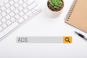 A white desk with a keyboard, a small plant, a notebook, and a search bar overlay that says "ADS."