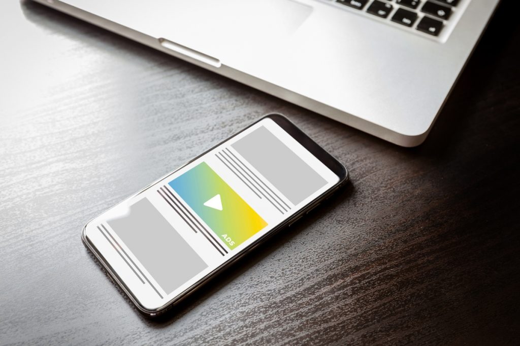 A smartphone displaying a video ad mockup lying on a dark wooden desk next to a laptop.