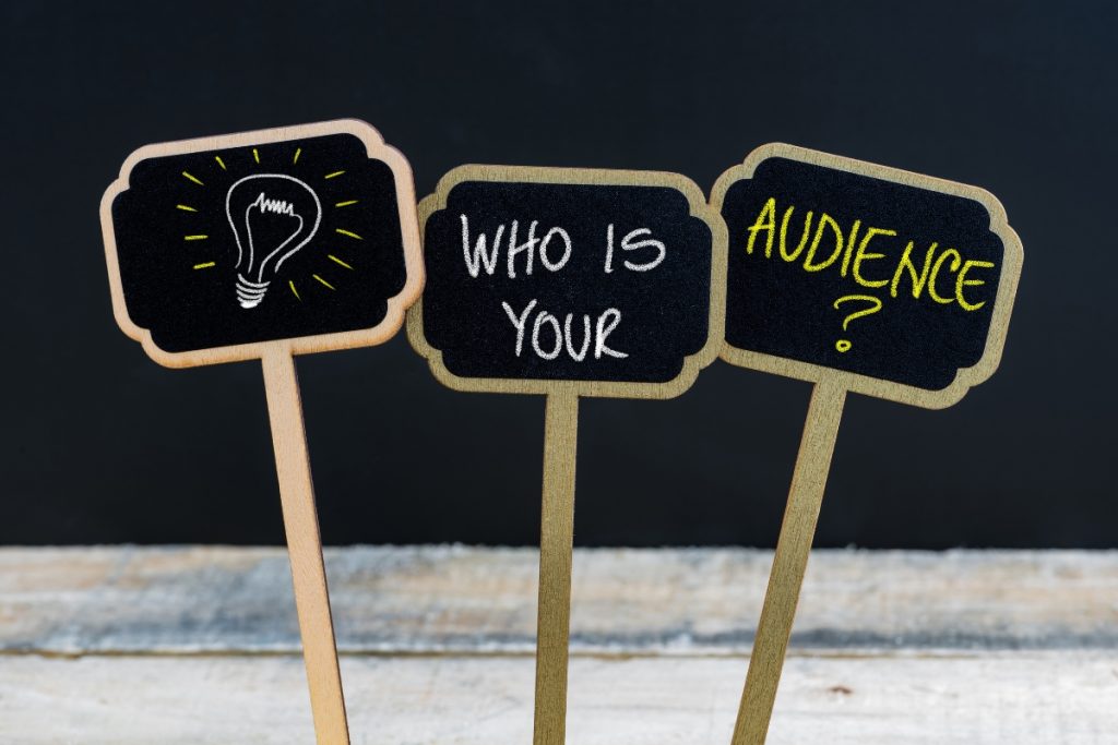 Three small handheld chalkboards on a wooden surface; the middle and right boards ask, "WHO IS YOUR AUDIENCE?"