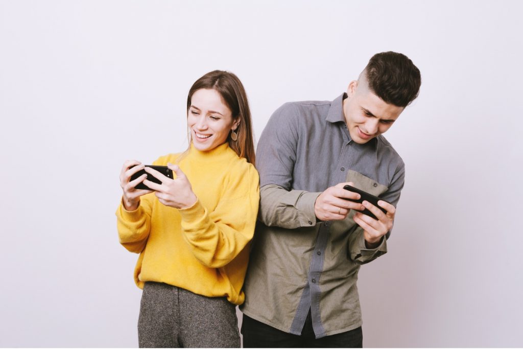 Two young adults standing back-to-back while both are focused on using their smartphones.
