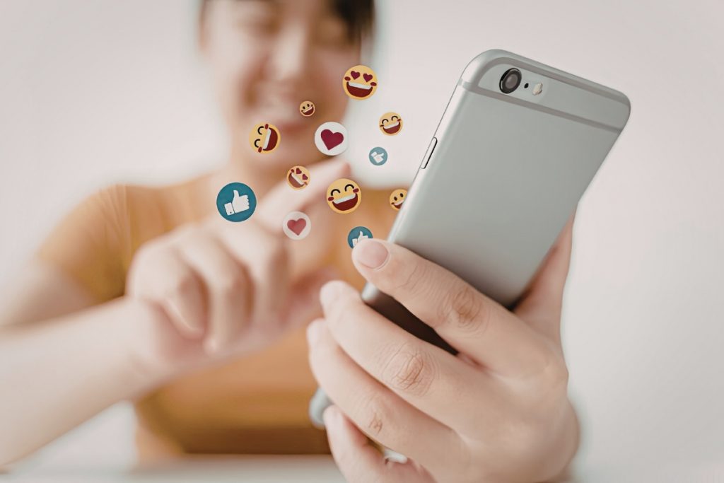 A close-up of a person's hands using a smartphone with various social media emojis floating above the screen.