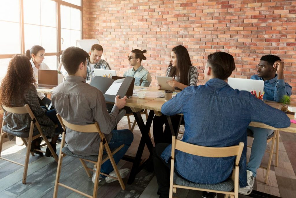 A diverse group of young professionals having a collaborative meeting around a wooden table in a modern office with a brick wall.