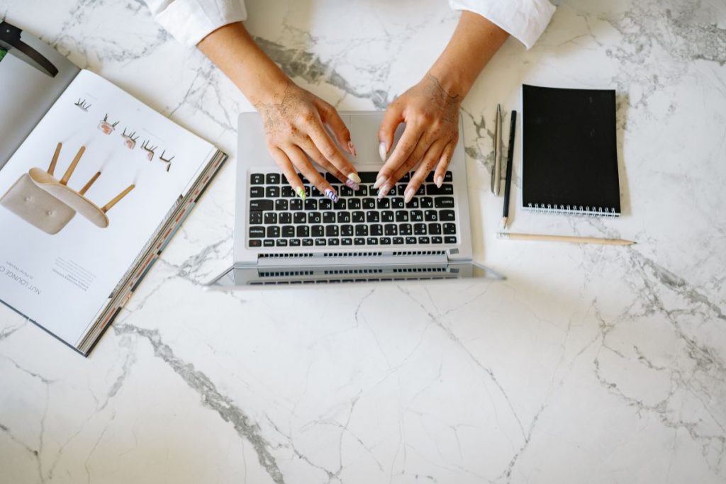 A person's hands typing on a laptop keyboard next to a notebook and a cup of coffee on a wooden desk.