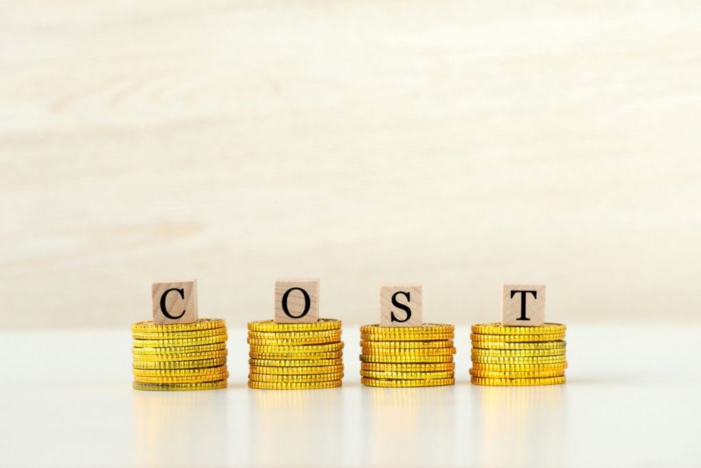 Four stacks of gold coins with wooden blocks on top that spell out the word "COST" against a light wooden background.
