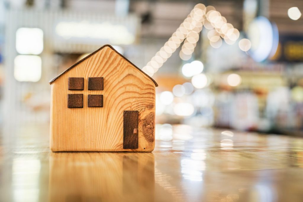 A small wooden toy house sitting on a reflective surface with a blurred background of warm indoor lights.