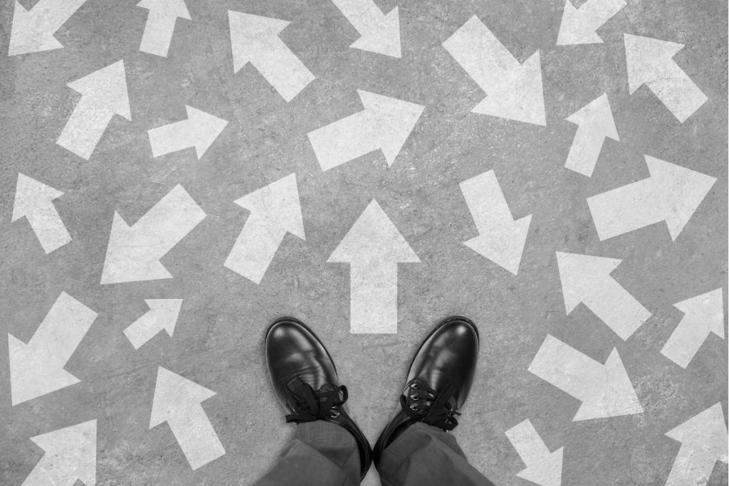 Top-down view of a person's black shoes standing on pavement covered in white arrows pointing in many different directions.