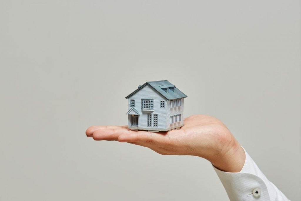 A person's hand holding a small, detailed model of a white two-story house against a plain grey background.