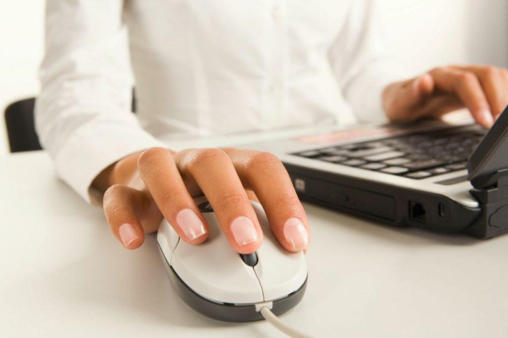 A close-up shot of a person's hand using a white computer mouse on a white desk, with a laptop open and hands typing in the background.