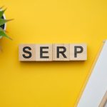 Four wooden toy blocks spelling out the acronym "SERP" on a bright yellow background, positioned next to a small green plant and a white spiral notebook with a pencil.