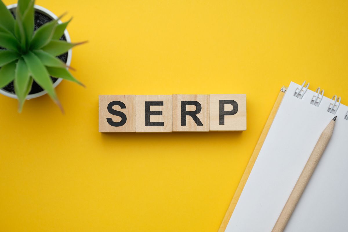 Four wooden toy blocks spelling out the acronym "SERP" on a bright yellow background, positioned next to a small green plant and a white spiral notebook with a pencil.