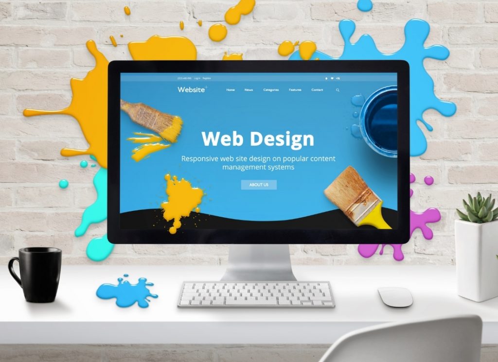 Desktop monitor showing a web design landing page against a brick wall with colourful paint splatters.