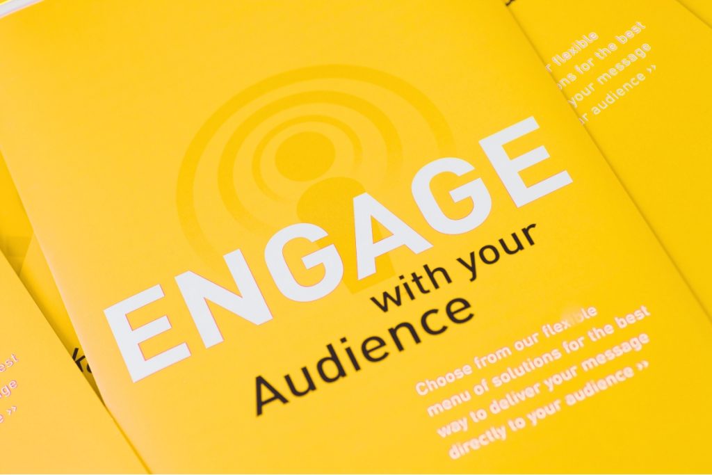 A close-up view of a bright yellow booklet featuring the bold white text "ENGAGE" and black text that reads "with your audience" alongside a subtle signal wave graphic.